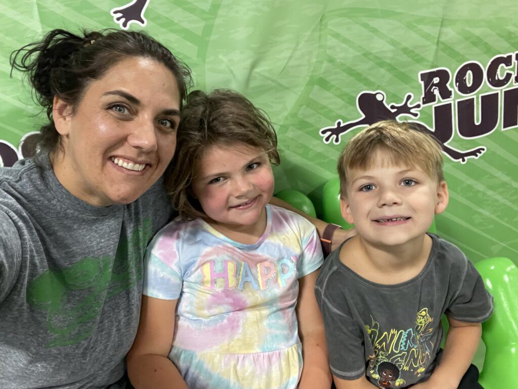 Woman smiling with two young children in front of a green Rock Jump backdrop.