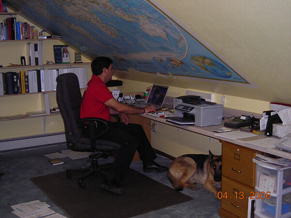 Man working at a computer in a small office with a dog resting beside his desk.